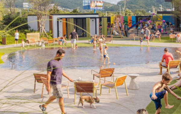 O Passeio Sapiens, novo empreendimento no norte da Ilha de Santa Catarina, será um centro de compras e de convivência a céu aberto. Na foto, o térreo do Passeio Primavera. Ao centro, um lago e um homem passeando com seu cão. Ao lado direito, crianças brincando em meio à grama verde.